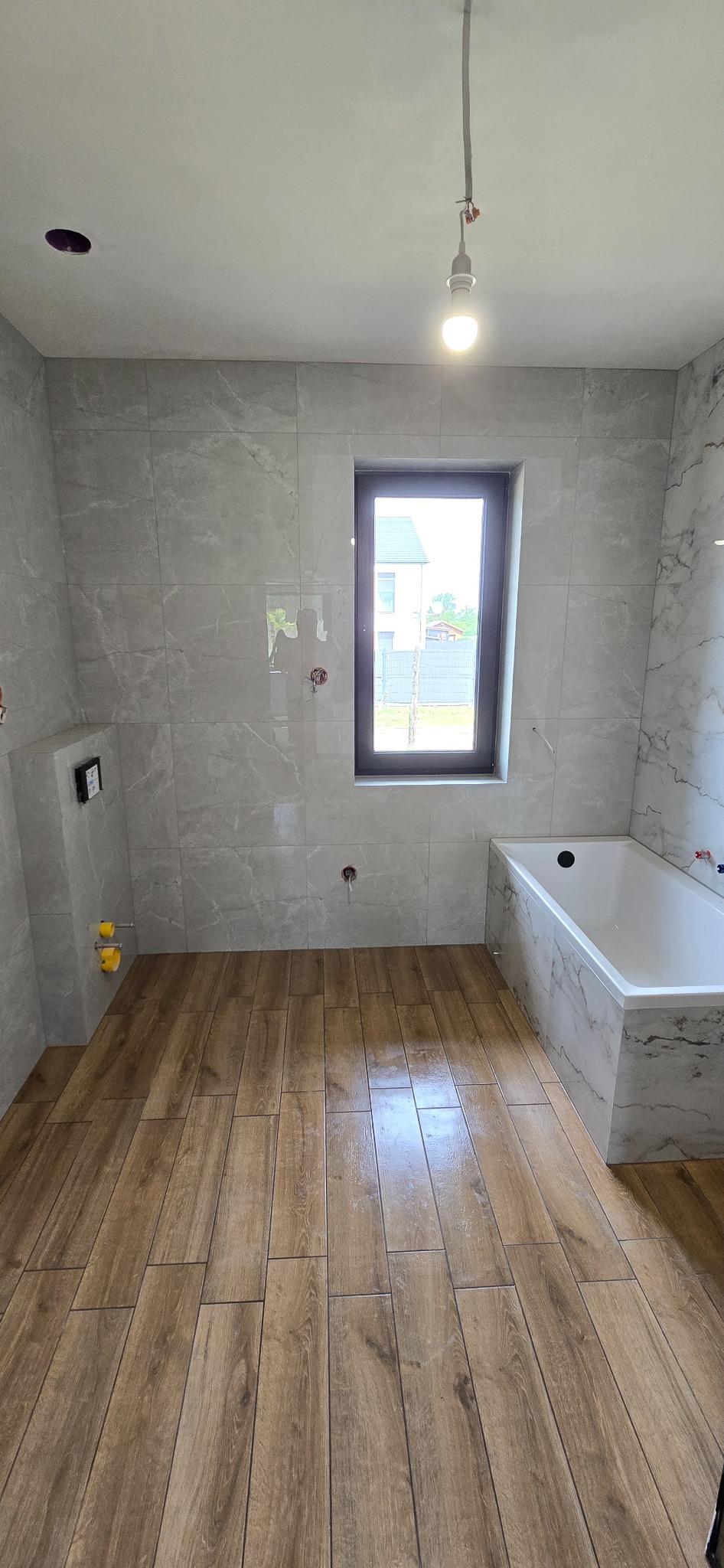 Wykończona łazienka z wanną, szarą marmurkową glazurą ścienną i drewnopodobną podłogą, widok na zewnątrz przez okno. Przewód żarówki zwisa z sufitu.