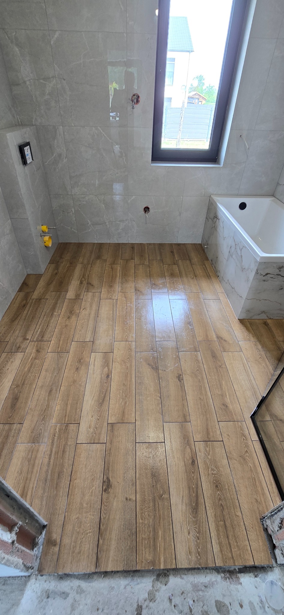 Ukończona podłoga z płytek imitujących drewno w łazience z oknem dachowym. Płytki ścienne w odcieniach szarości z delikatnym wzorem marmuru.