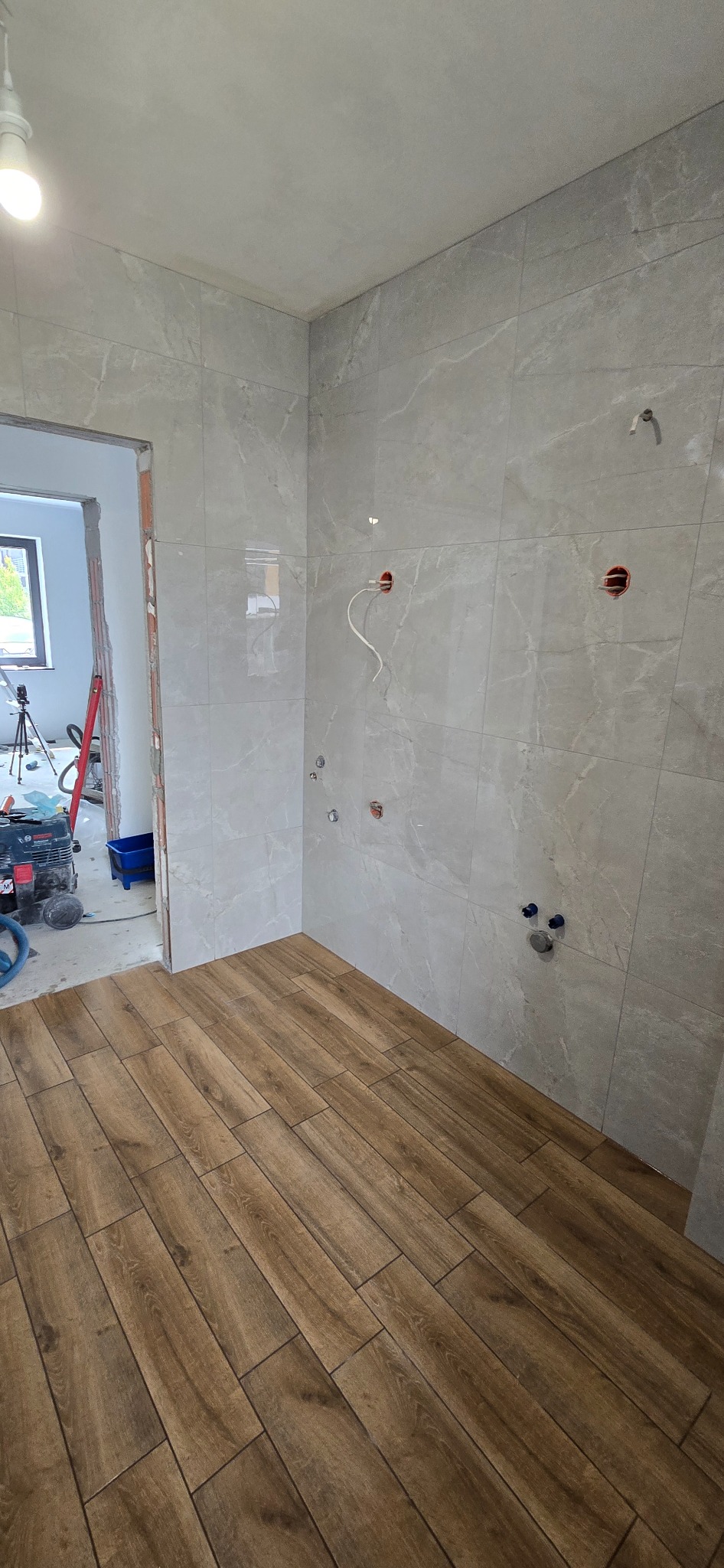 Wykończona łazienka z szarymi płytkami imitującymi marmur na ścianach i drewnopodobną podłogą. Widoczne instalacje wodne i elektryczne.