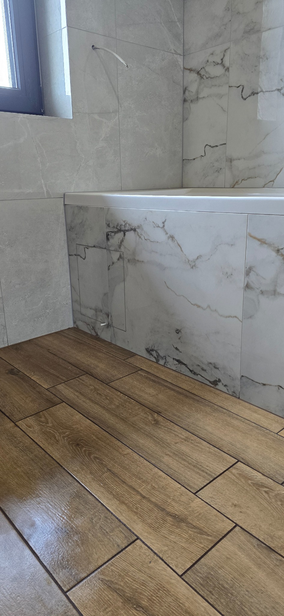 Wykończona łazienka: brązowe płytki imitujące drewno na podłodze, marmurowe płytki na ścianach i obudowie wanny. Widoczny fragment okna.