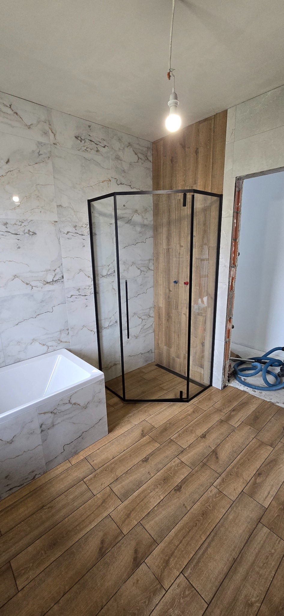 Wykończona łazienka z kabiną prysznicową typu walk-in z czarnymi profilami, drewnopodobną podłogą i marmurowymi płytkami ściennymi. Widoczna wanna i surowy otwór drzwiowy.