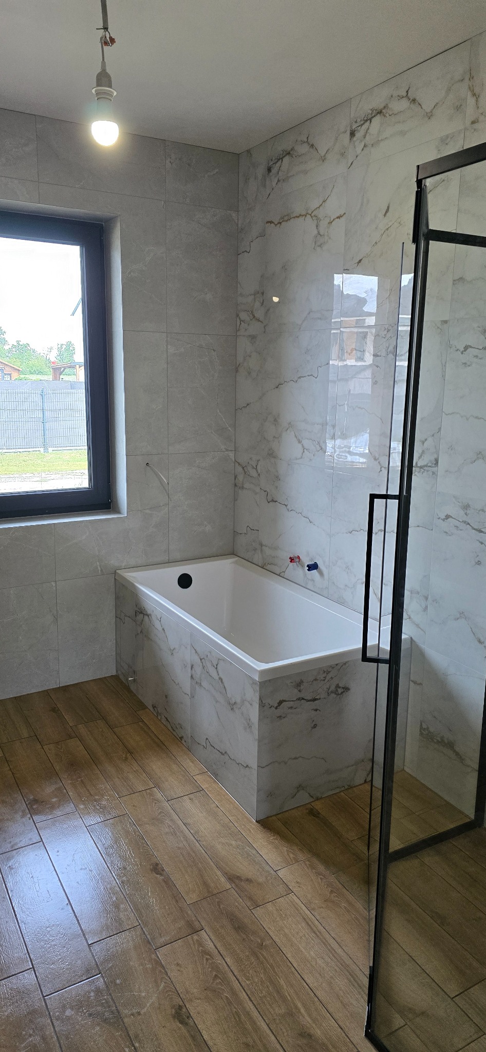 Nowoczesna łazienka z wanną obudowaną płytkami imitującymi marmur, drewnopodobna podłoga i czarna rama kabiny prysznicowej. Widok na okno.