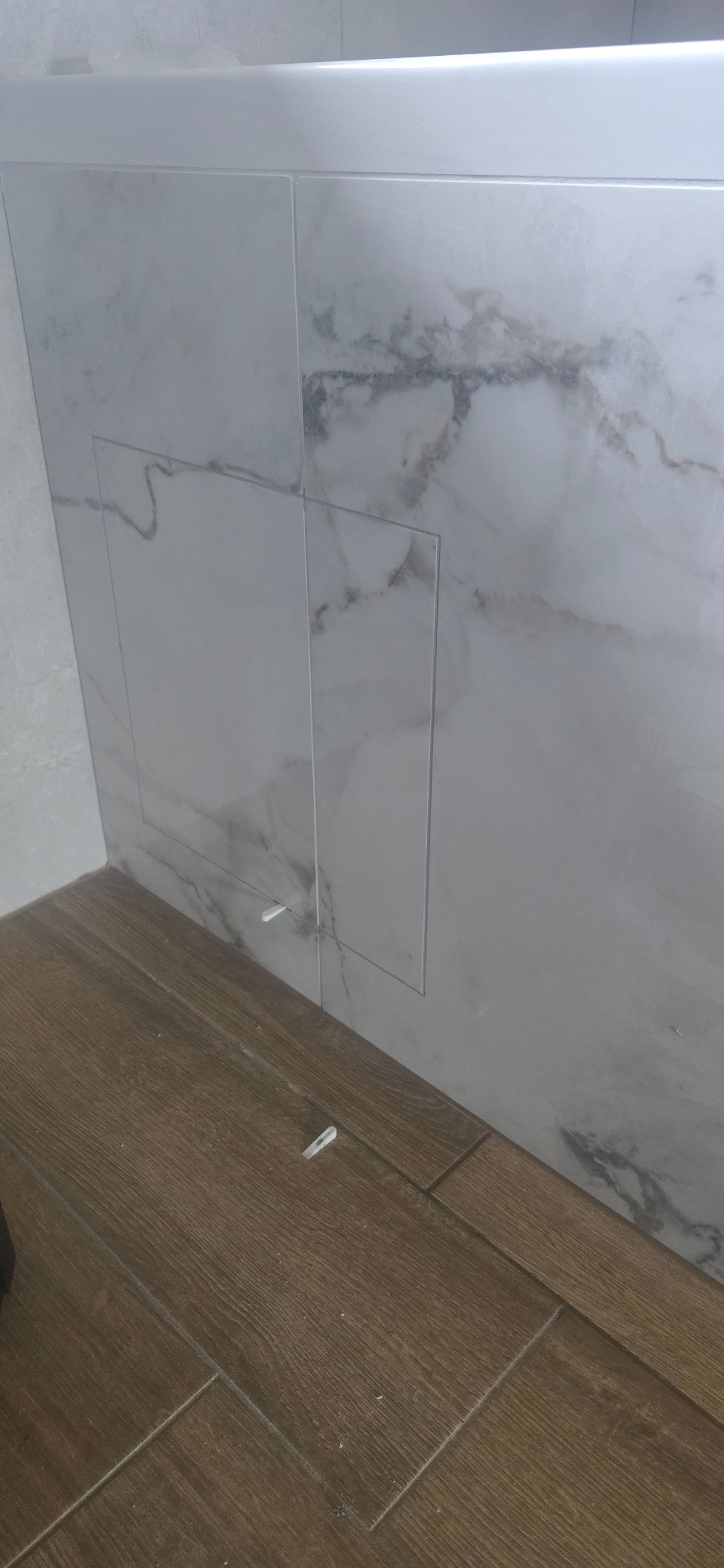 Wykończona ściana z płytek imitujących marmur z rewizją, widoczne płytki podłogowe imitujące drewno i krzyżyki dystansowe.