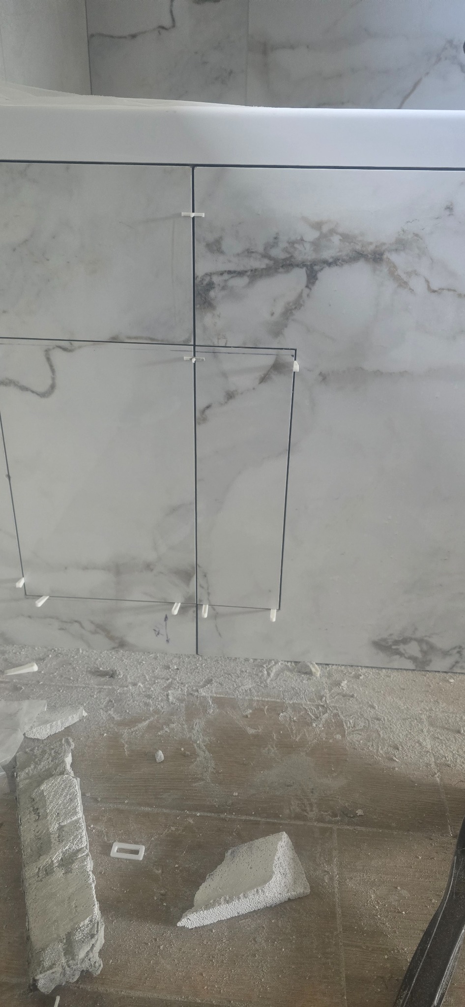 Montaż płytek imitujących marmur na ścianie, widoczne krzyżyki dystansowe, fragment podłogi z resztkami zaprawy. Praca w toku, zbliżenie na fragment ściany.