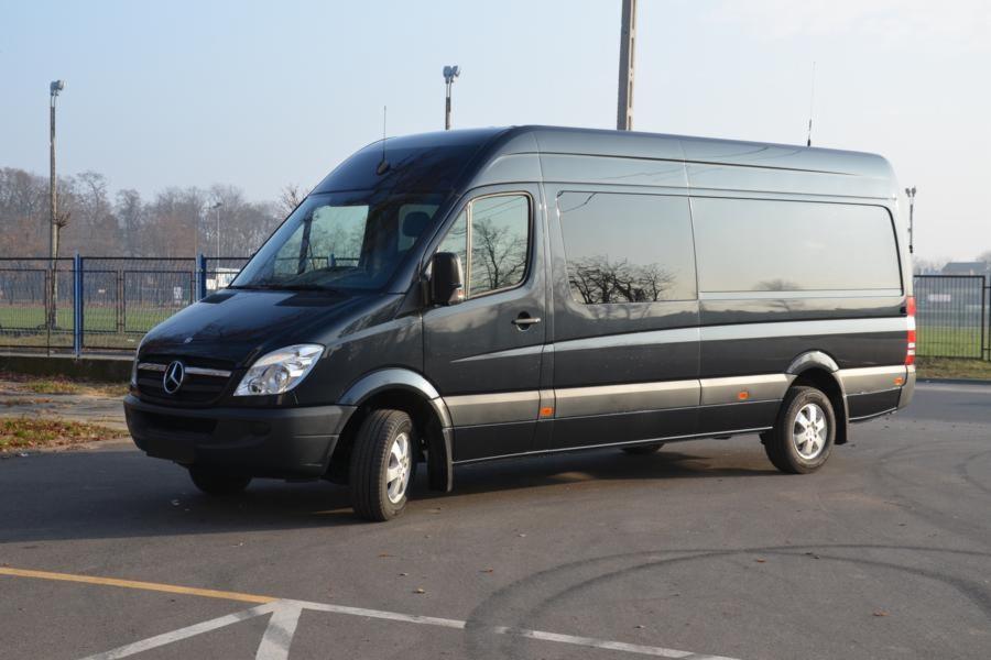 Czarny Mercedes Sprinter z długim rozstawem osi, zaparkowany na asfaltowym placu z widocznymi śladami opon, w tle ogrodzenie i drzewa.
