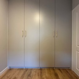Piotr Wojtoń - Czterodrzwiowa szafa wnękowa w odcieniach szarości z podłużnymi złotymi uchwytami, dopasowana do koloru ścian, z widoczną częścią drewnianej podłogi i białych drzwi po prawej stronie.