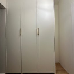 Piotr Wojtoń - Trzydrzwiowa, biała szafa wnękowa z podłużnymi, złotymi uchwytami, dopasowana do wnętrza z drewnianą podłogą.