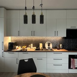 Piotr Wojtoń - Nowoczesna, jasna kuchnia z białymi szafkami, czarnymi uchwytami i trzema wiszącymi lampami z czarnymi kloszami. Na blacie widoczne sprzęty AGD i akcesoria kuchenne.