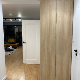 Piotr Wojtoń - Wysoka szafa wnękowa z drewnianymi frontami, wkomponowana w nowoczesne wnętrze z drewnianą podłogą i widokiem na salon. Minimalistyczny design, jasne oświetlenie.
