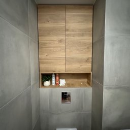 Piotr Wojtoń - Wnękowa szafka łazienkowa z drewnianymi frontami, wkomponowana w szare płytki. Na półce dekoracje: roślina, kosmetyki i papier toaletowy. Widoczny fragment toalety i pralki.