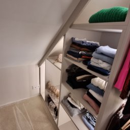 Piotr Wojtoń - Szafa wnękowa na poddaszu ze skośnym sufitem, półki wypełnione ubraniami, butami i torebką. Jasne wnętrze, dywan w tle.