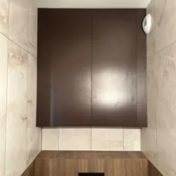 Nowoczesna, brązowa szafka łazienkowa nad zabudową WC, z wentylatorem. Jasne, marmurowe płytki na ścianach. Minimalistyczny design.