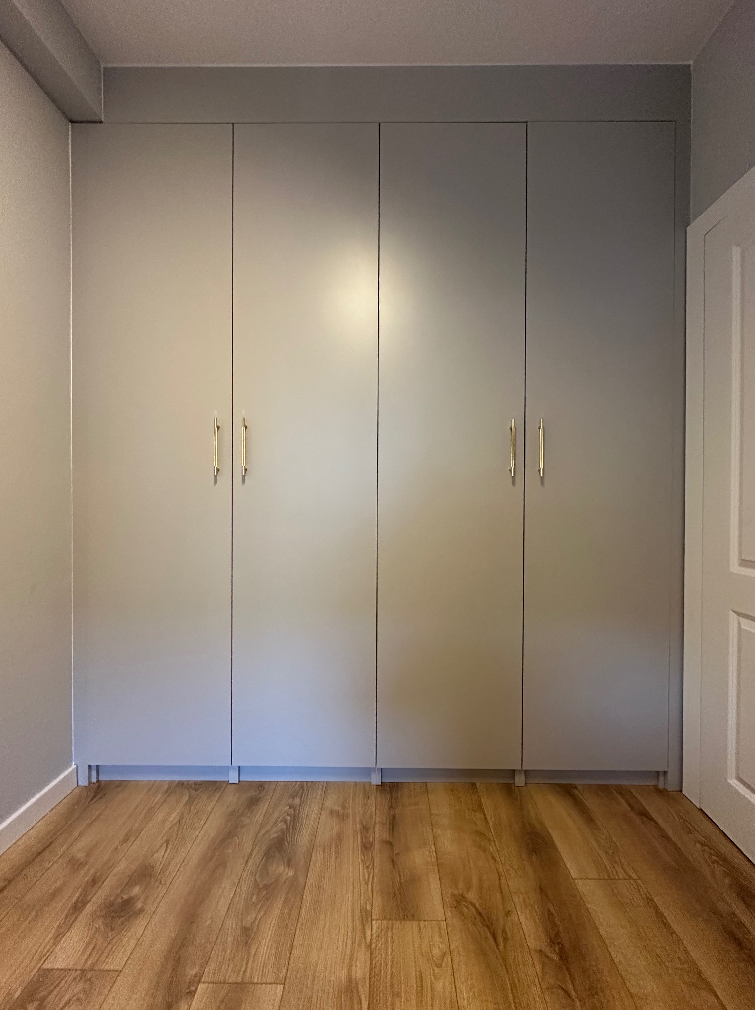 Czterodrzwiowa szafa wnękowa w odcieniach szarości z podłużnymi złotymi uchwytami, dopasowana do koloru ścian, z widoczną częścią drewnianej podłogi i białych drzwi po prawej stronie.