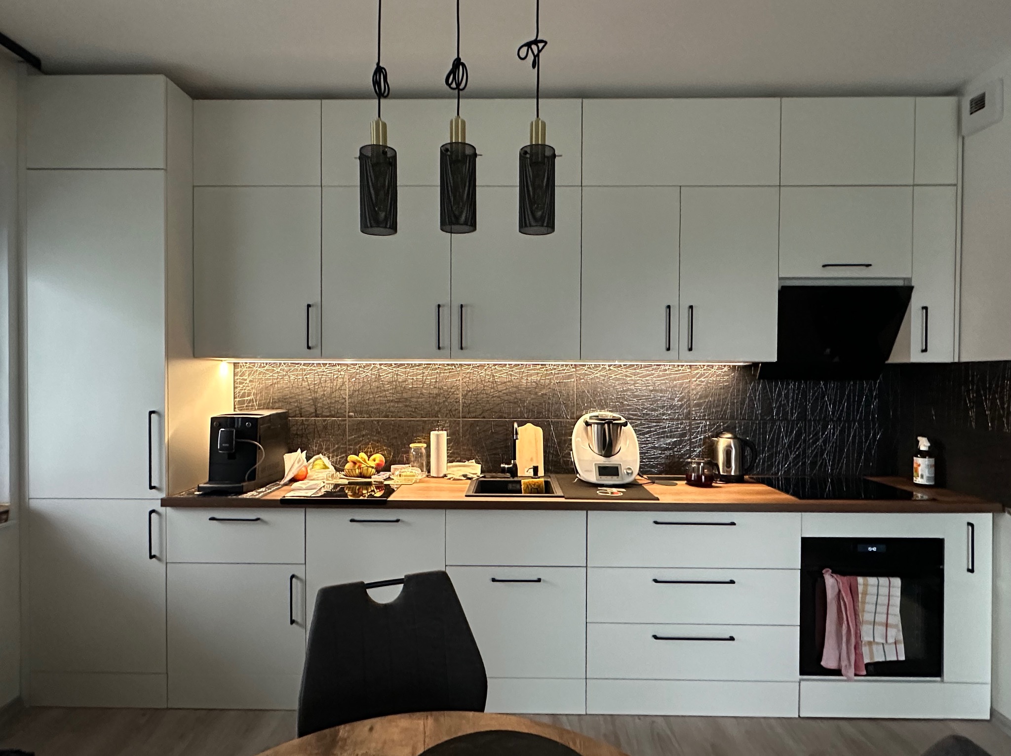 Nowoczesna, jasna kuchnia z białymi szafkami, czarnymi uchwytami i trzema wiszącymi lampami z czarnymi kloszami. Na blacie widoczne sprzęty AGD i akcesoria kuchenne.