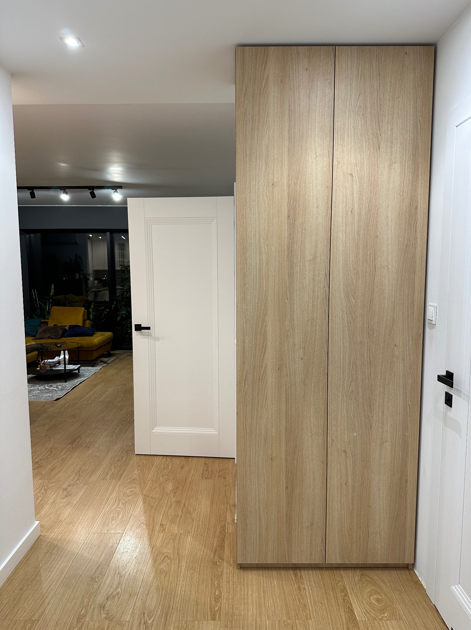 Wysoka szafa wnękowa z drewnianymi frontami, wkomponowana w nowoczesne wnętrze z drewnianą podłogą i widokiem na salon. Minimalistyczny design, jasne oświetlenie.