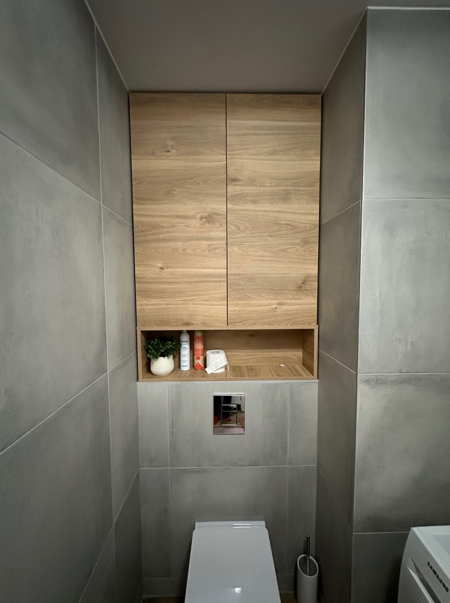 Wnękowa szafka łazienkowa z drewnianymi frontami, wkomponowana w szare płytki. Na półce dekoracje: roślina, kosmetyki i papier toaletowy. Widoczny fragment toalety i pralki.