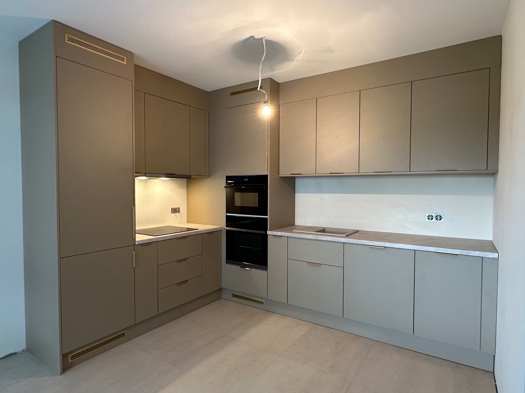 Nowoczesna kuchnia w odcieniach beżu z minimalistycznymi uchwytami i subtelnym oświetleniem, prezentująca zabudowę na wymiar z piekarnikiem w zabudowie.