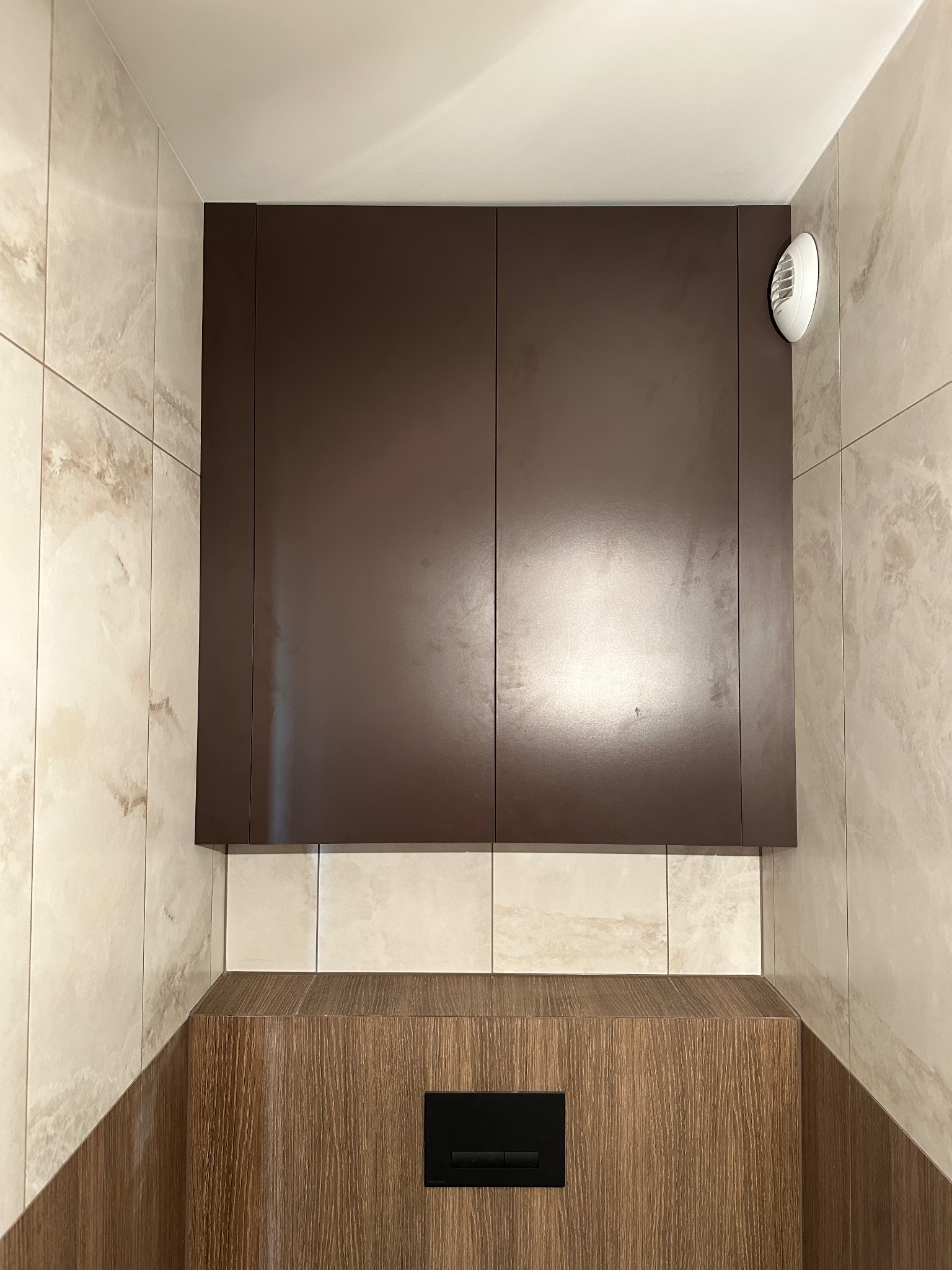 Nowoczesna, brązowa szafka łazienkowa nad zabudową WC, z wentylatorem. Jasne, marmurowe płytki na ścianach. Minimalistyczny design.
