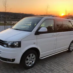Biały Volkswagen Transporter T6 z relingami dachowymi i progami, zaparkowany na brukowanej powierzchni, widok z przodu, zachód słońca w tle.