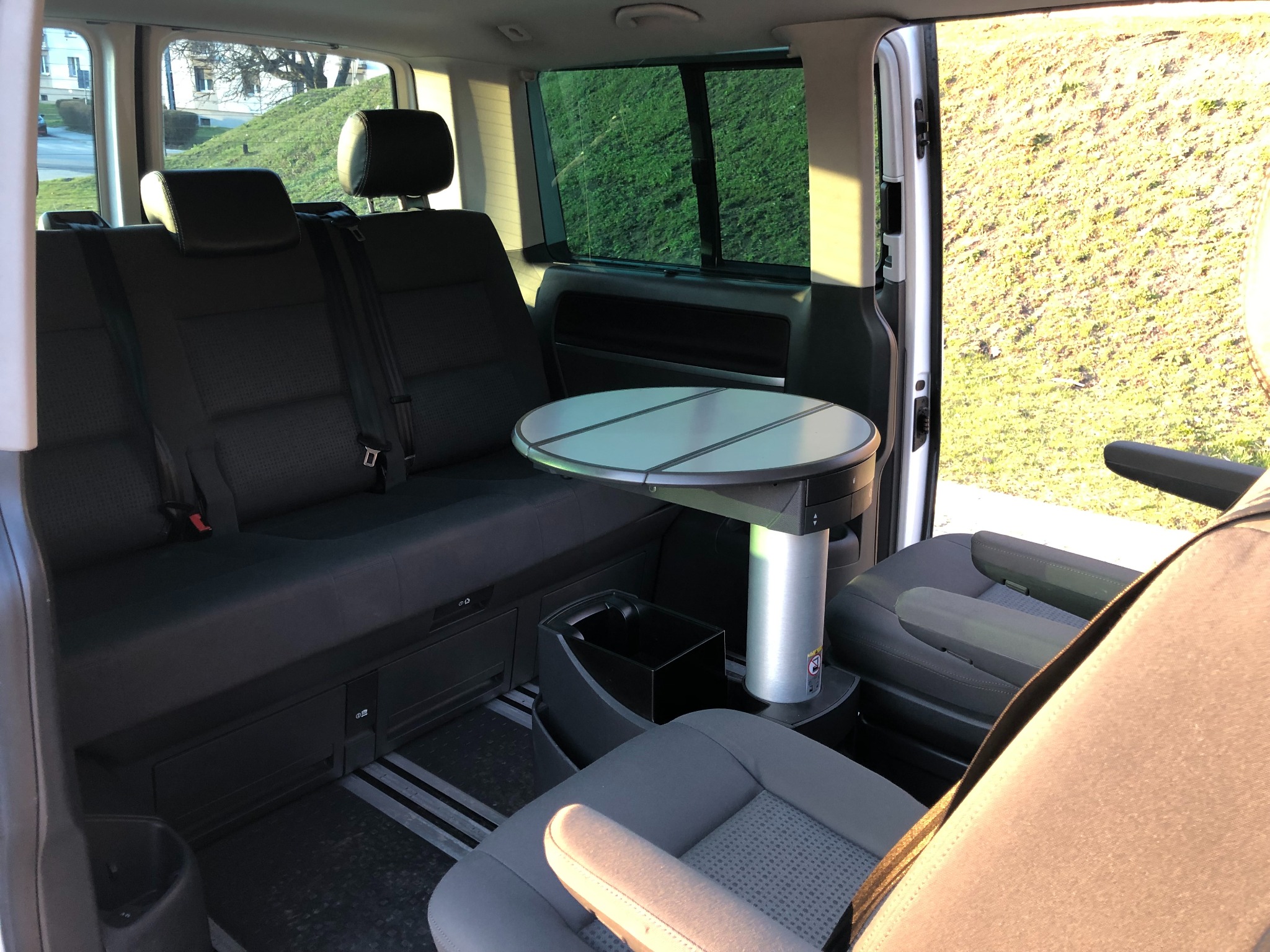 Wnętrze białego busa z czarnymi siedzeniami, rozkładanym okrągłym stolikiem i widokiem na zielony trawnik za oknem.