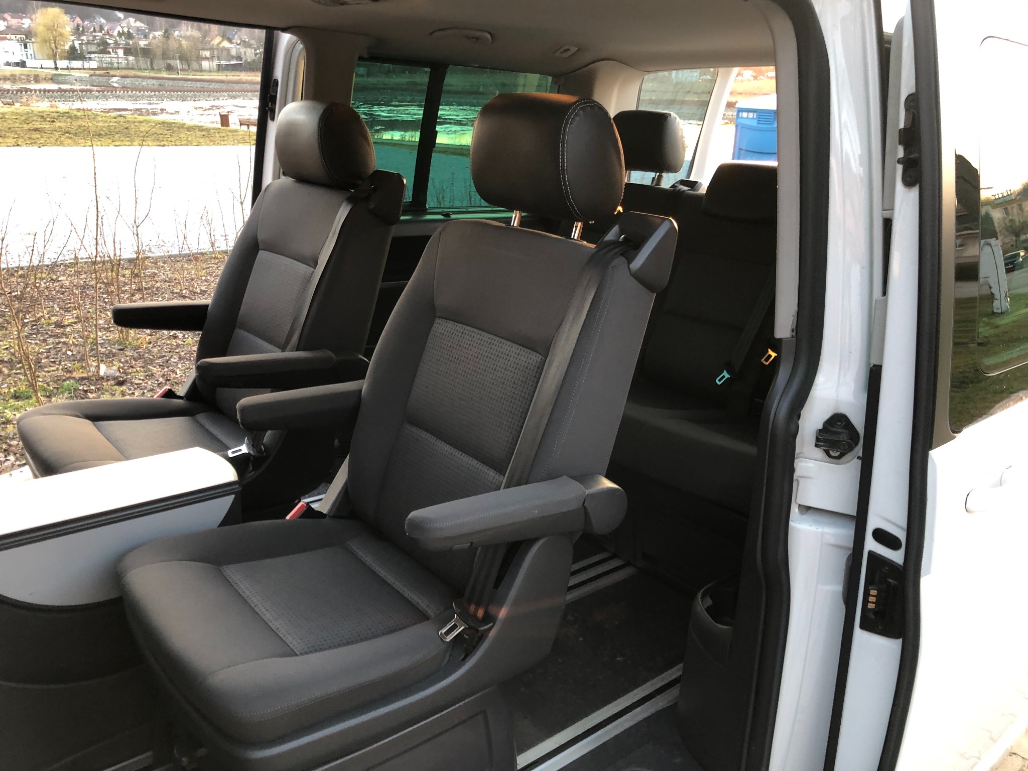 Wnętrze białego busa z trzema rzędami szarych foteli z regulowanymi podłokietnikami, widok z otwartych drzwi na rzekę w tle.