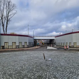 Kleszczów: Widok na plac z kostki brukowej z krzesłem na pierwszym planie, w tle budynek w trakcie budowy z widocznymi elementami konstrukcyjnymi i przejazdem. Na dalszym planie ciężki sprzęt...