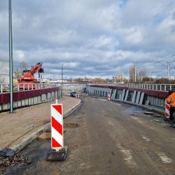 WMR-BUDOWNICTWO Mariusz Rutkowski - Ujęcie z budowy wiaduktu z widoczną czerwoną żurawiem Manitou, pracownikiem w pomarańczowej kamizelce odblaskowej ustawiającym biało-czerwone bariery ostrzegawcze, oraz ułożonymi płytami chodnikowymi.