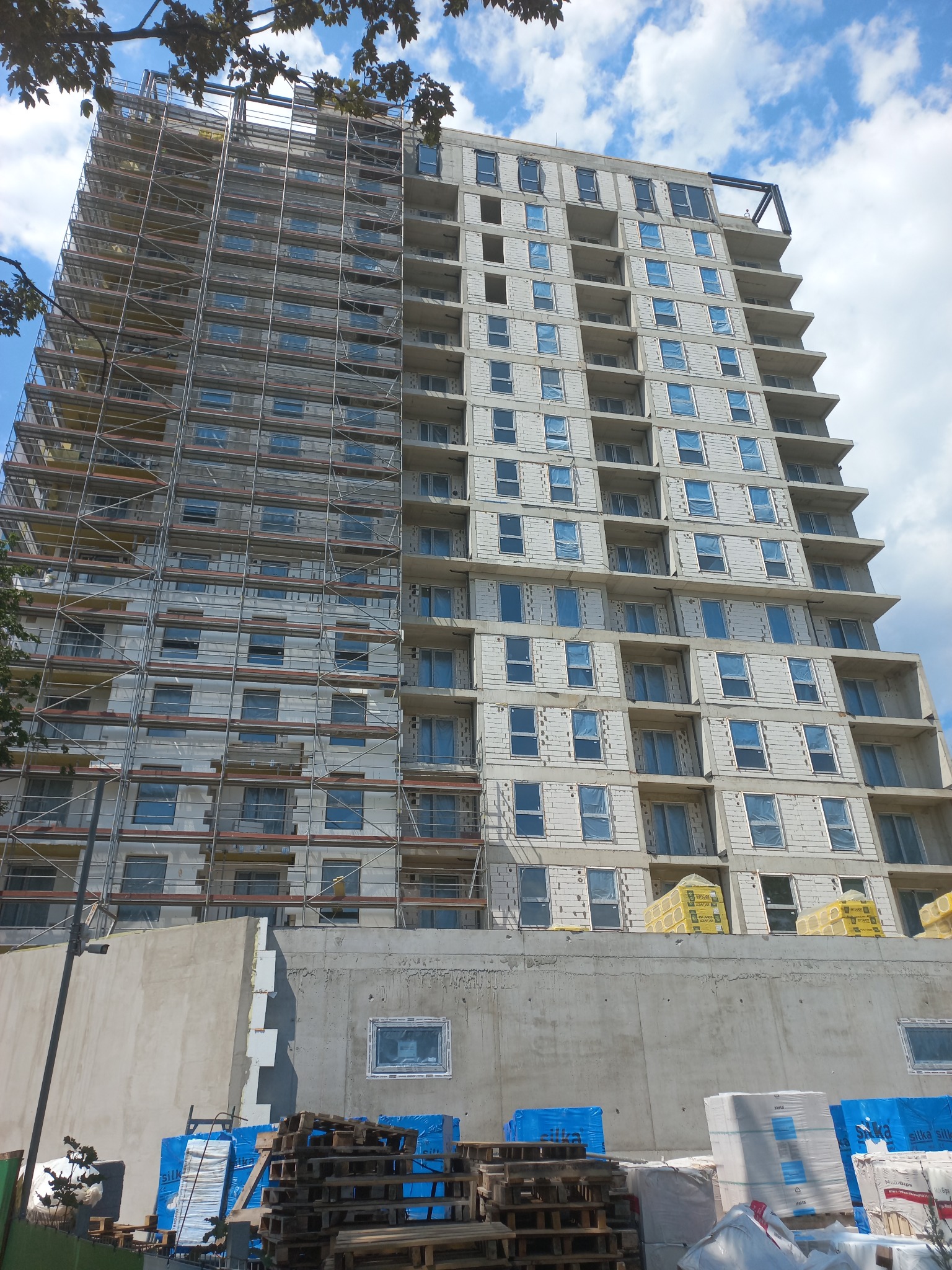 Wysoki budynek w trakcie budowy, z widocznymi rusztowaniami po jednej stronie i oknami zabezpieczonymi folią po drugiej, z materiałami budowlanymi składowanymi na paletach u podstawy