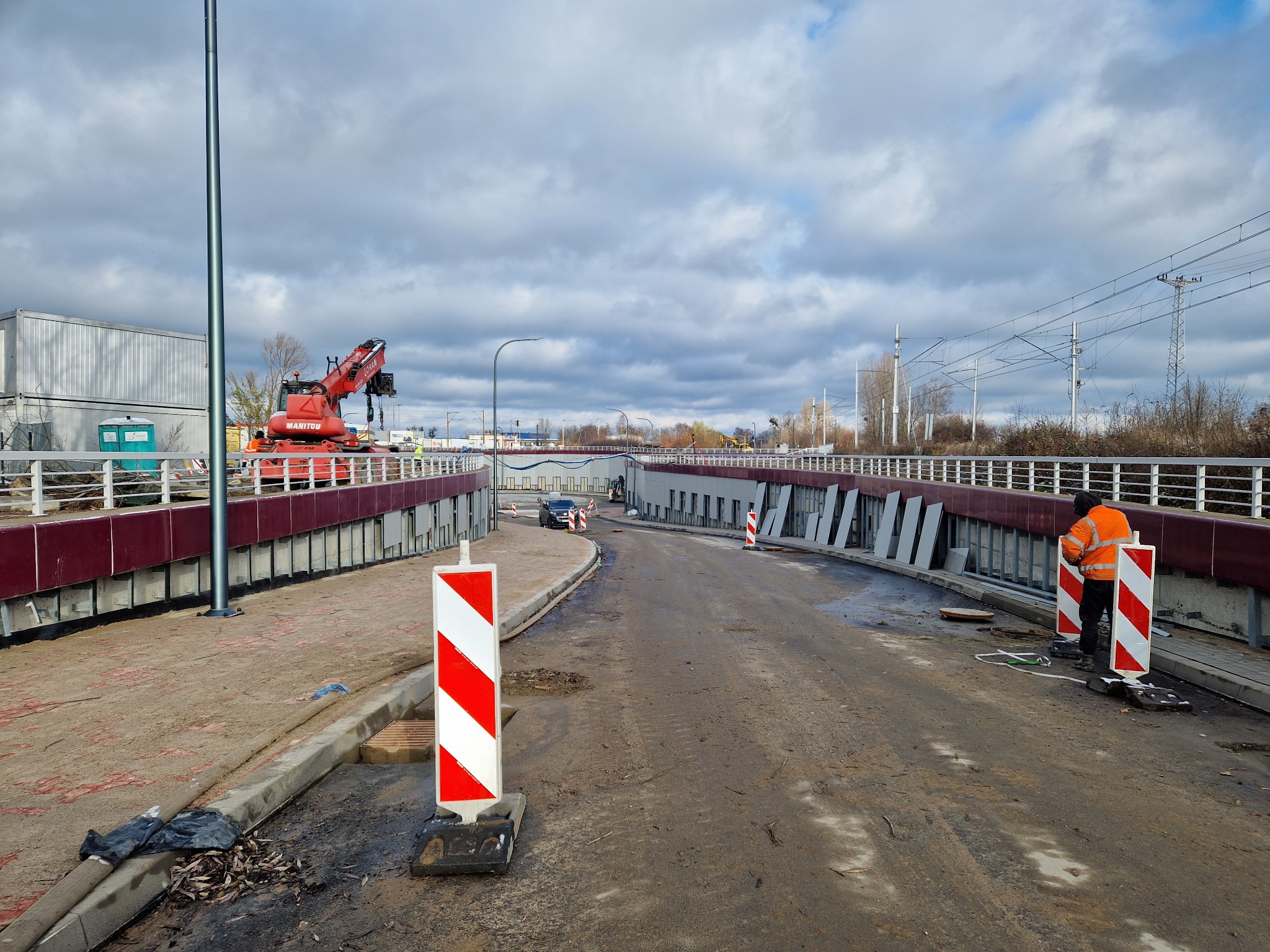Ujęcie z budowy wiaduktu z widoczną czerwoną żurawiem Manitou, pracownikiem w pomarańczowej kamizelce odblaskowej ustawiającym biało-czerwone bariery ostrzegawcze, oraz ułożonymi płytami chodnikowymi.