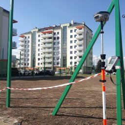 Usługi geodezyjne inż. Marek Cisek - GEOGRUNT Rzeszów - Pomiar geodezyjny na terenie budowy nowego osiedla mieszkaniowego, widoczny odbiornik GNSS na tyczce, otoczony zielonymi słupkami i taśmą ostrzegawczą, budynek mieszkalny w tle.