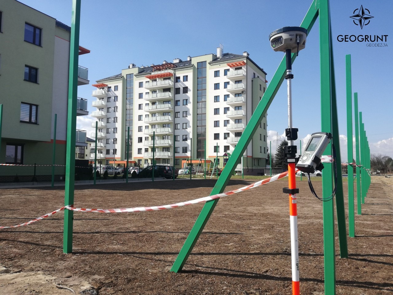 Pomiar geodezyjny na terenie budowy nowego osiedla mieszkaniowego, widoczny odbiornik GNSS na tyczce, otoczony zielonymi słupkami i taśmą ostrzegawczą, budynek mieszkalny w tle.