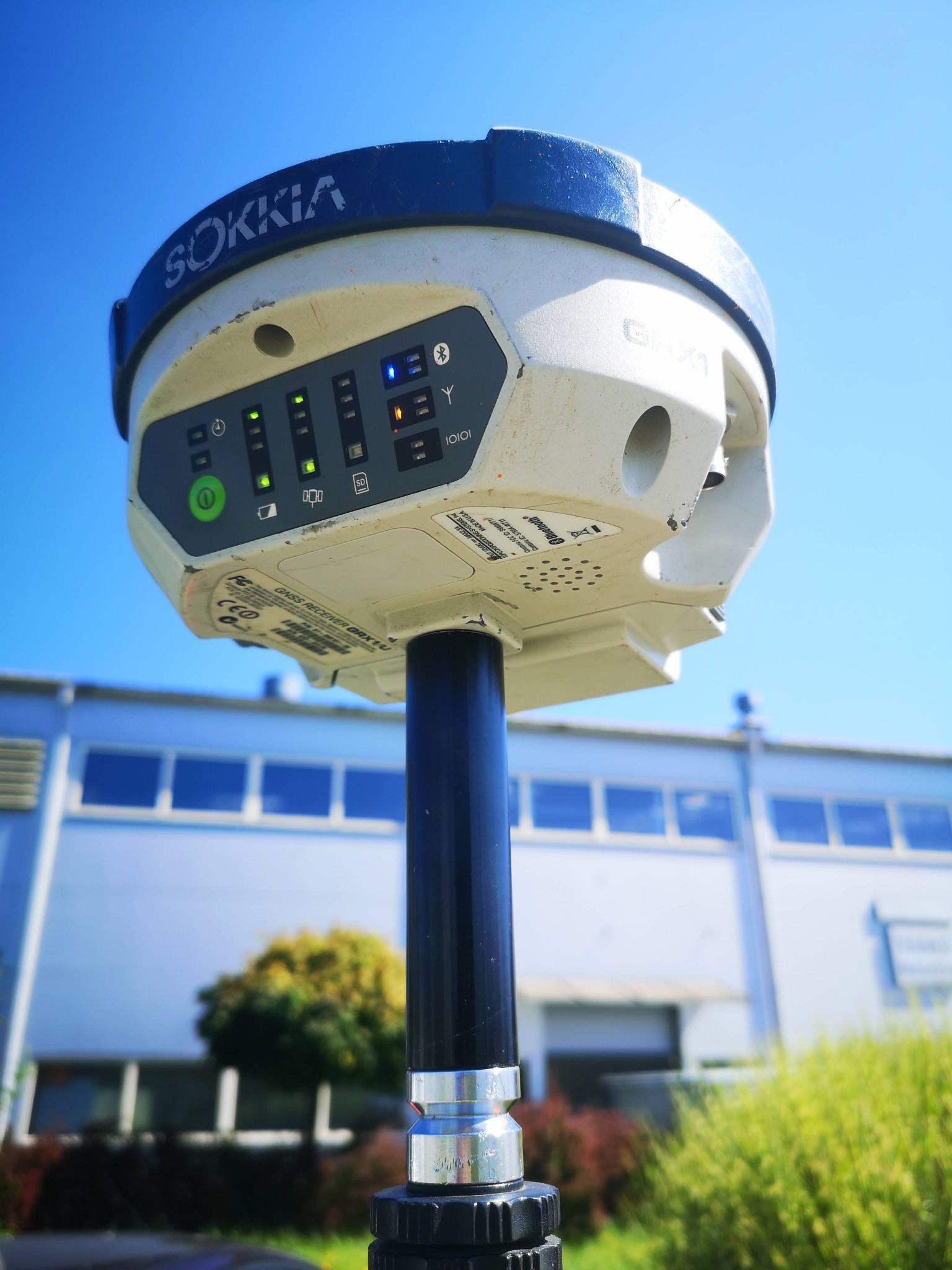 Geodezyjny odbiornik GNSS Sokkia GRX1U zamontowany na tyczce, z widocznymi diodami sygnalizacyjnymi, na tle błękitnego nieba i budynku.