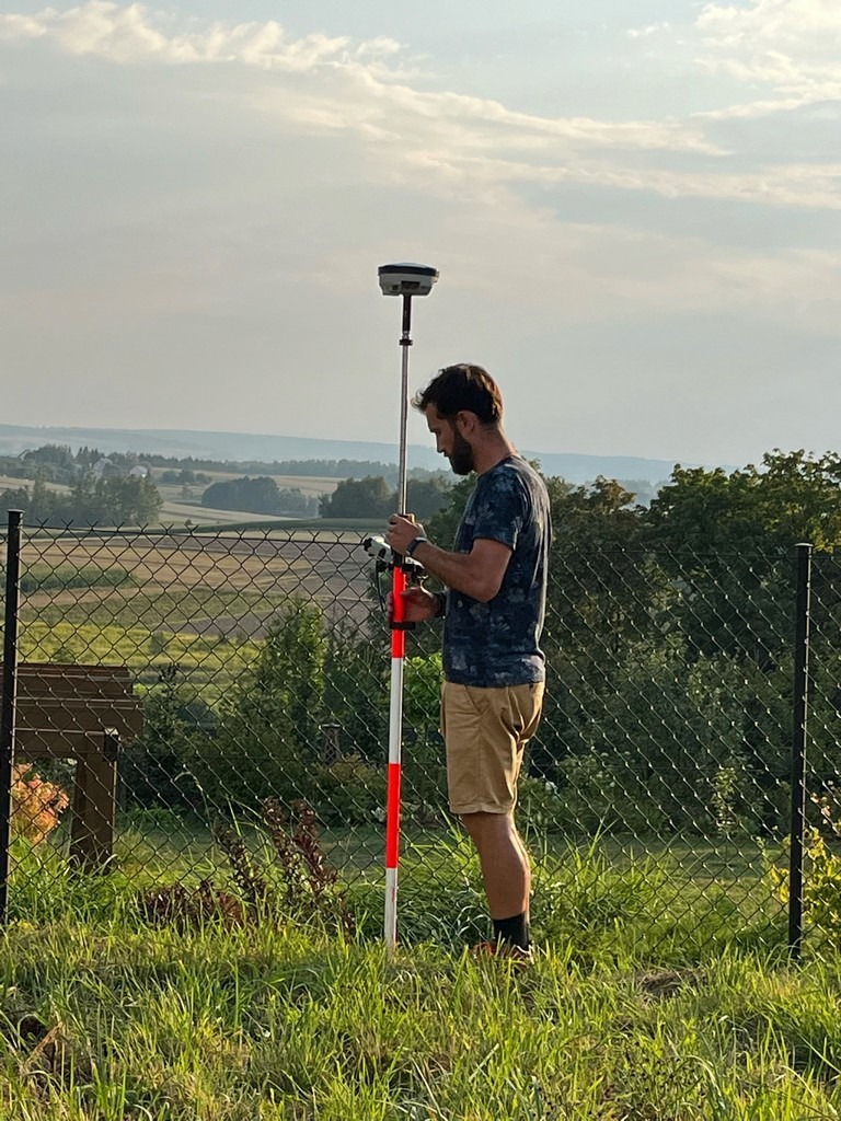 Mężczyzna w koszulce moro i beżowych szortach obsługuje geodezyjny odbiornik GPS na tyczce, na tle pagórkowatego krajobrazu za metalową siatką ogrodzeniową.
