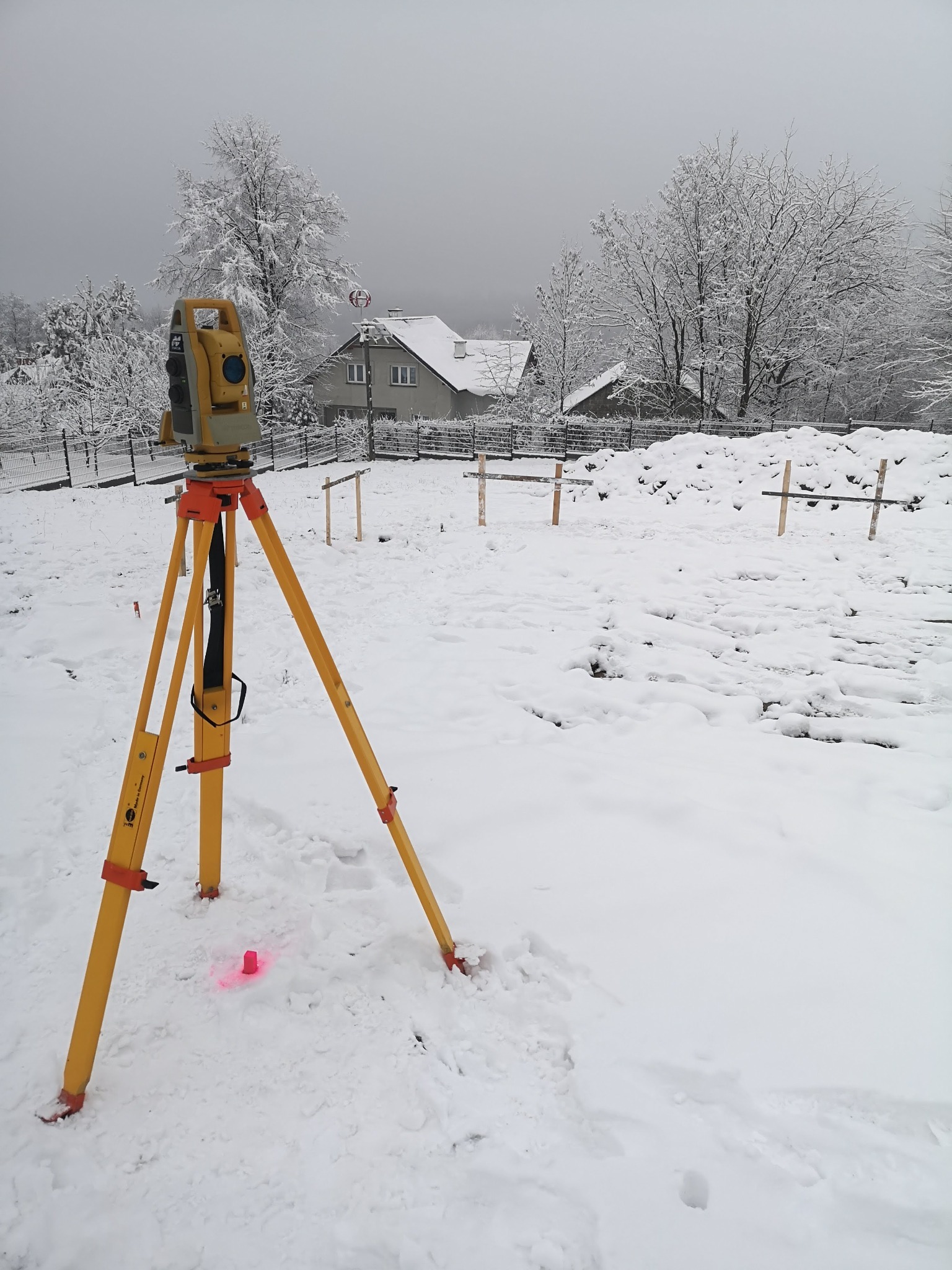 Żółty teodolit ustawiony na trójnogu na zasypanym śniegiem terenie budowy, w tle ośnieżone drzewa i domy, widoczny czerwony znacznik geodezyjny.
