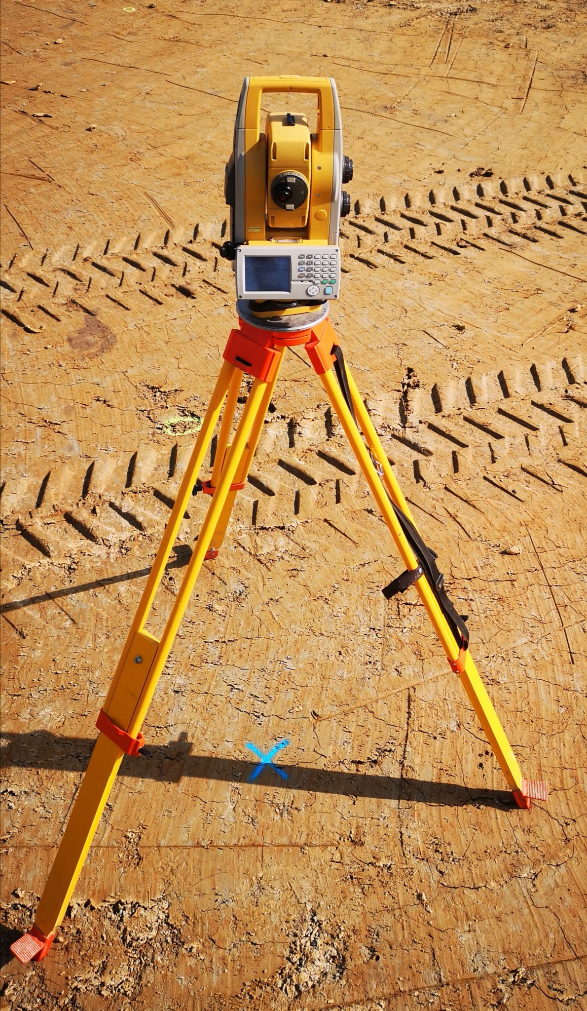 Żółty teodolit na trójnogu ustawiony na nierównym, gliniastym terenie, z wyraźnymi śladami opon i niebieskim oznaczeniem krzyżykiem.