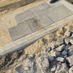 Usługi Brukarskie Rafał Duras - Etap prac: układanie szarej kostki brukowej na podsypce piaskowej, widoczne obrzeża i częściowo ułożone elementy. Osoba w czarnych butach stoi na obrzeżu.