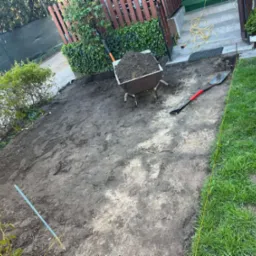 Teren przygotowany pod prace brukarskie: wytyczone granice żółtą linką, ziemia wyrównana, taczka z ziemią, łopata. Etap prac ziemnych przed ułożeniem kostki.