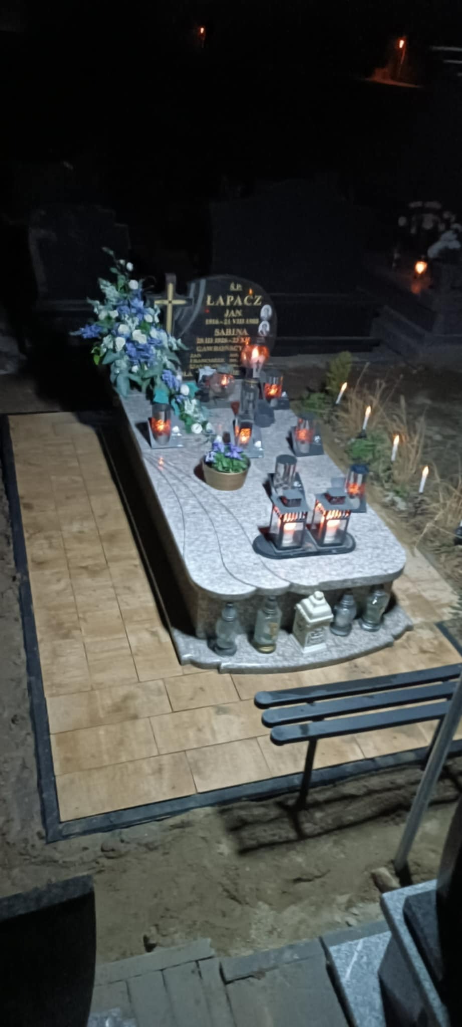 Oświetlony nagrobek z szarego granitu, otoczony kostką brukową w ciepłych odcieniach, kwiaty i znicze tworzą wieczorny klimat zadumy.