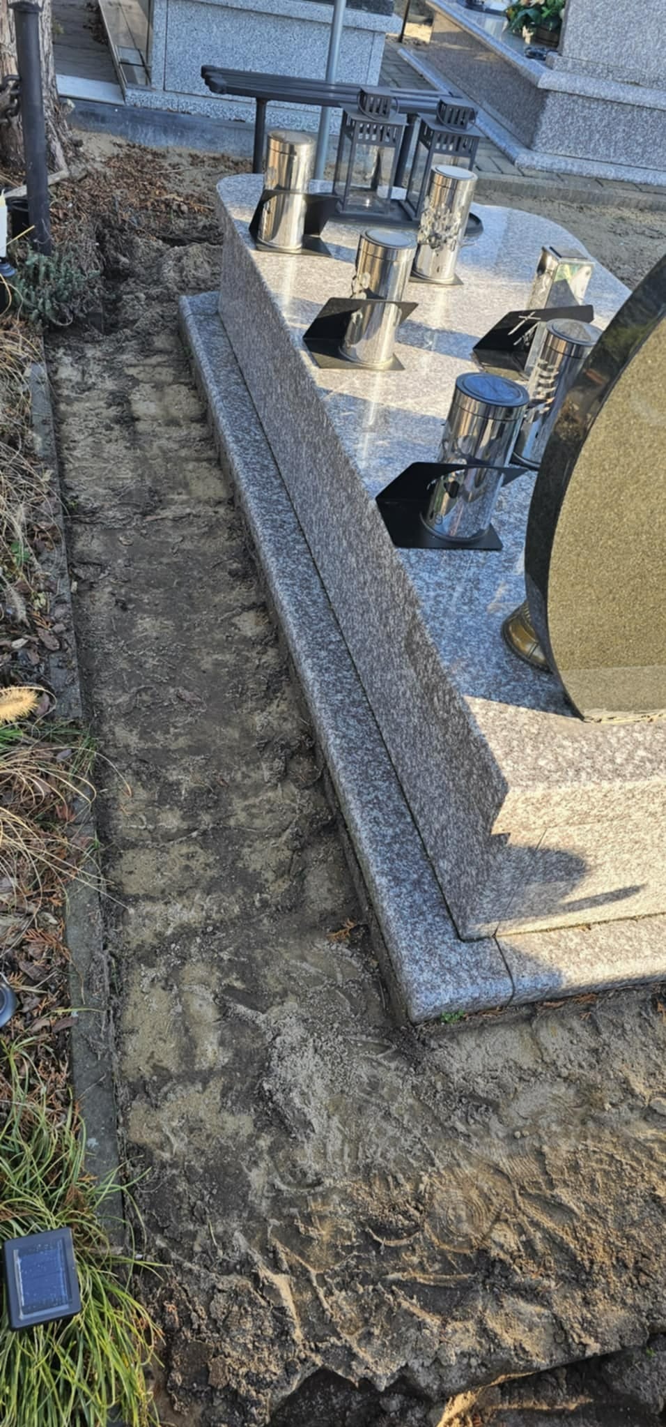 Przygotowanie terenu pod montaż obrzeży granitowych wokół nagrobka z szarego granitu, widoczna wykopana ziemia i ślady po pracach ziemnych.