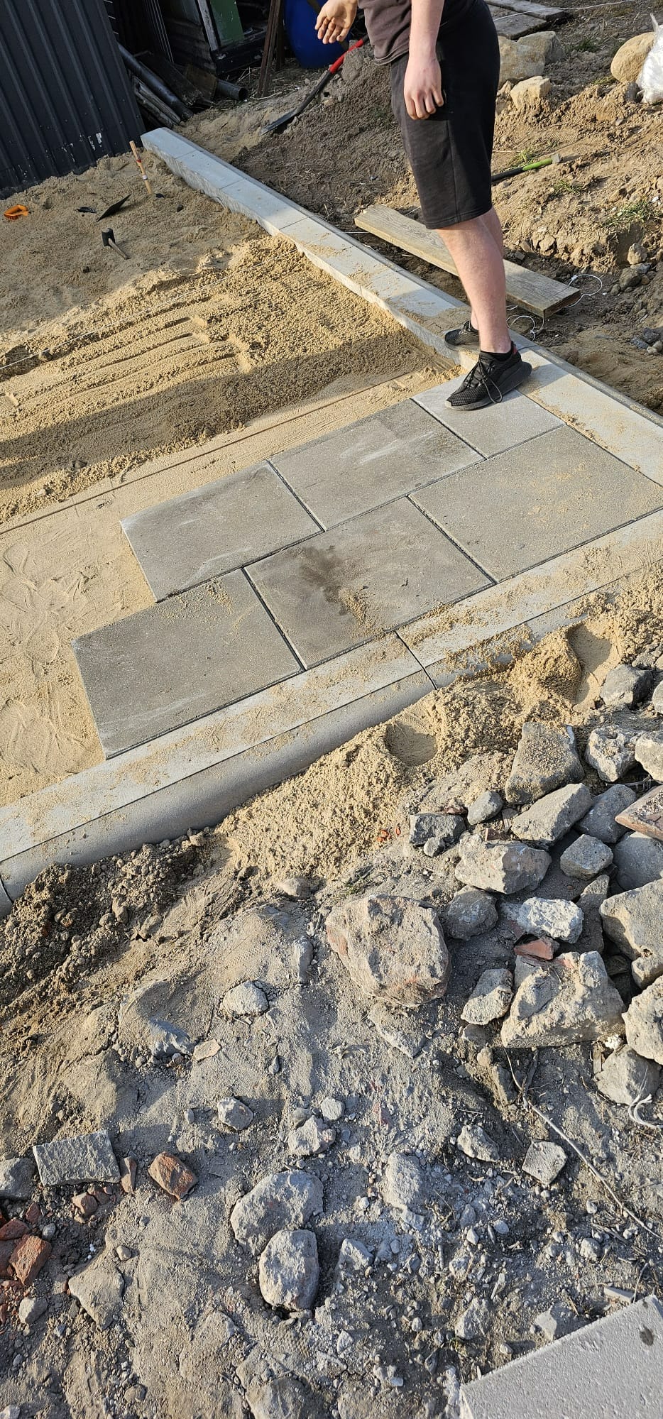 Etap prac: układanie szarej kostki brukowej na podsypce piaskowej, widoczne obrzeża i częściowo ułożone elementy. Osoba w czarnych butach stoi na obrzeżu.