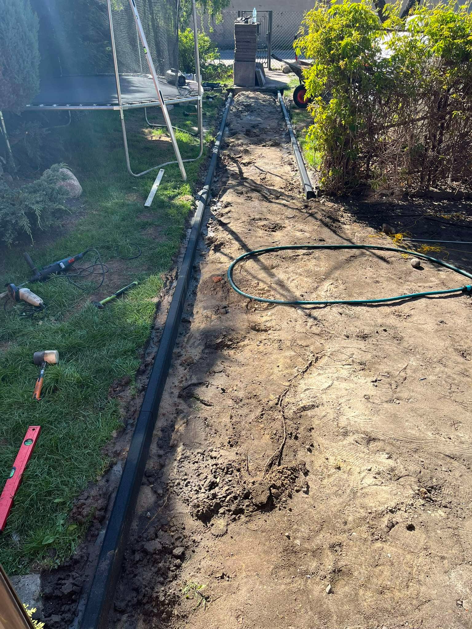 Przygotowanie podłoża: ułożone obrzeża z kostki, narzędzia pracy (poziomica, młotek, szlifierka) na trawie, widok na wytyczoną ścieżkę w ogrodzie.