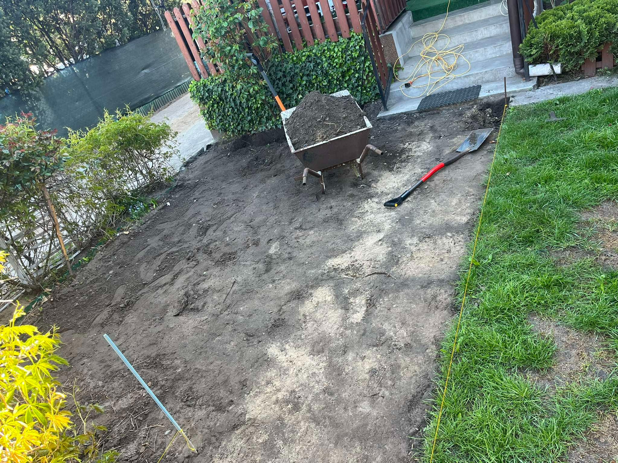 Teren przygotowany pod prace brukarskie: wytyczone granice żółtą linką, ziemia wyrównana, taczka z ziemią, łopata. Etap prac ziemnych przed ułożeniem kostki.