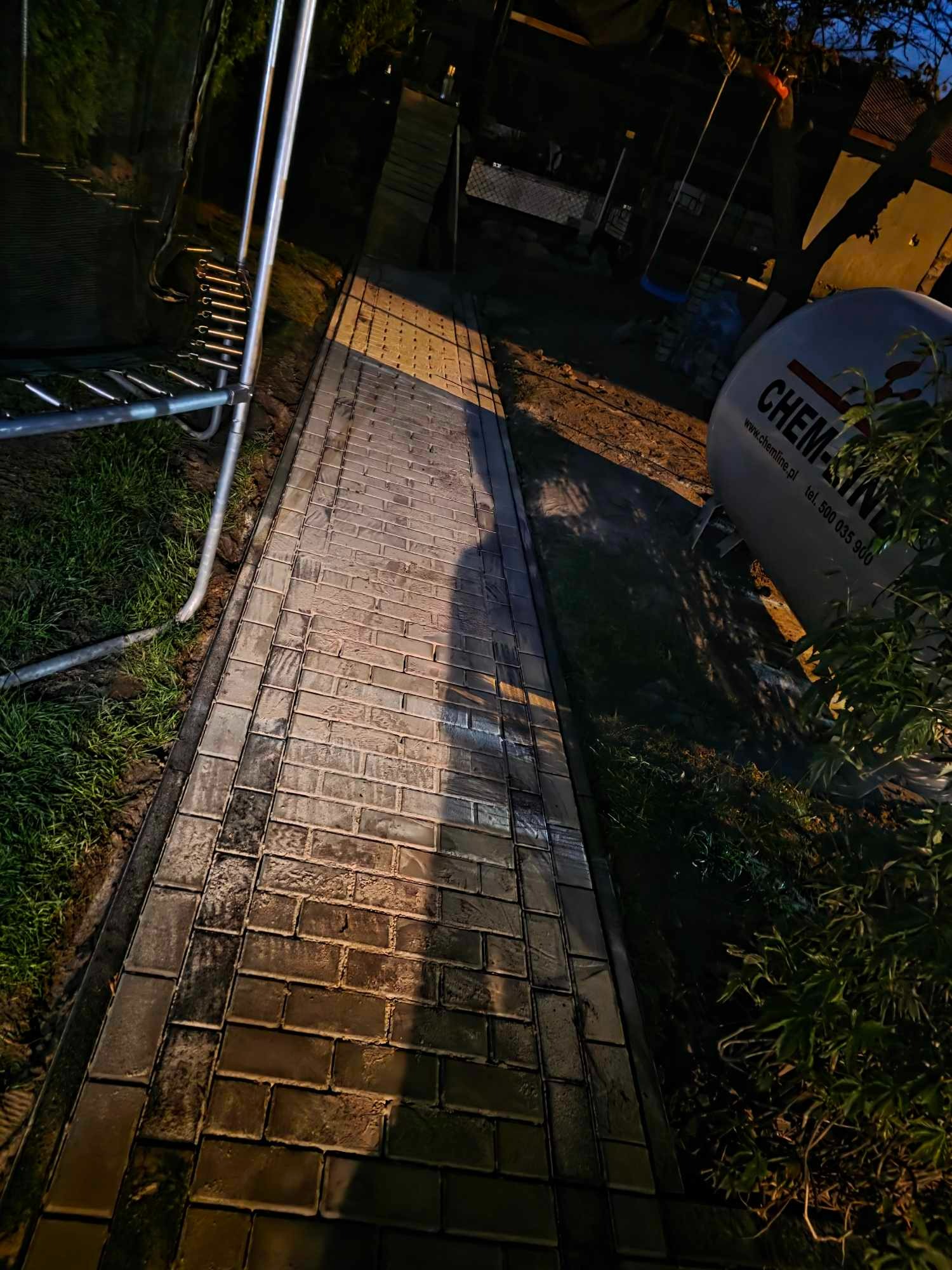 Świeżo ułożony chodnik z kostki brukowej w odcieniach szarości, oświetlony wieczornym światłem, z widocznym sprzętem budowlanym i fragmentem zieleni.