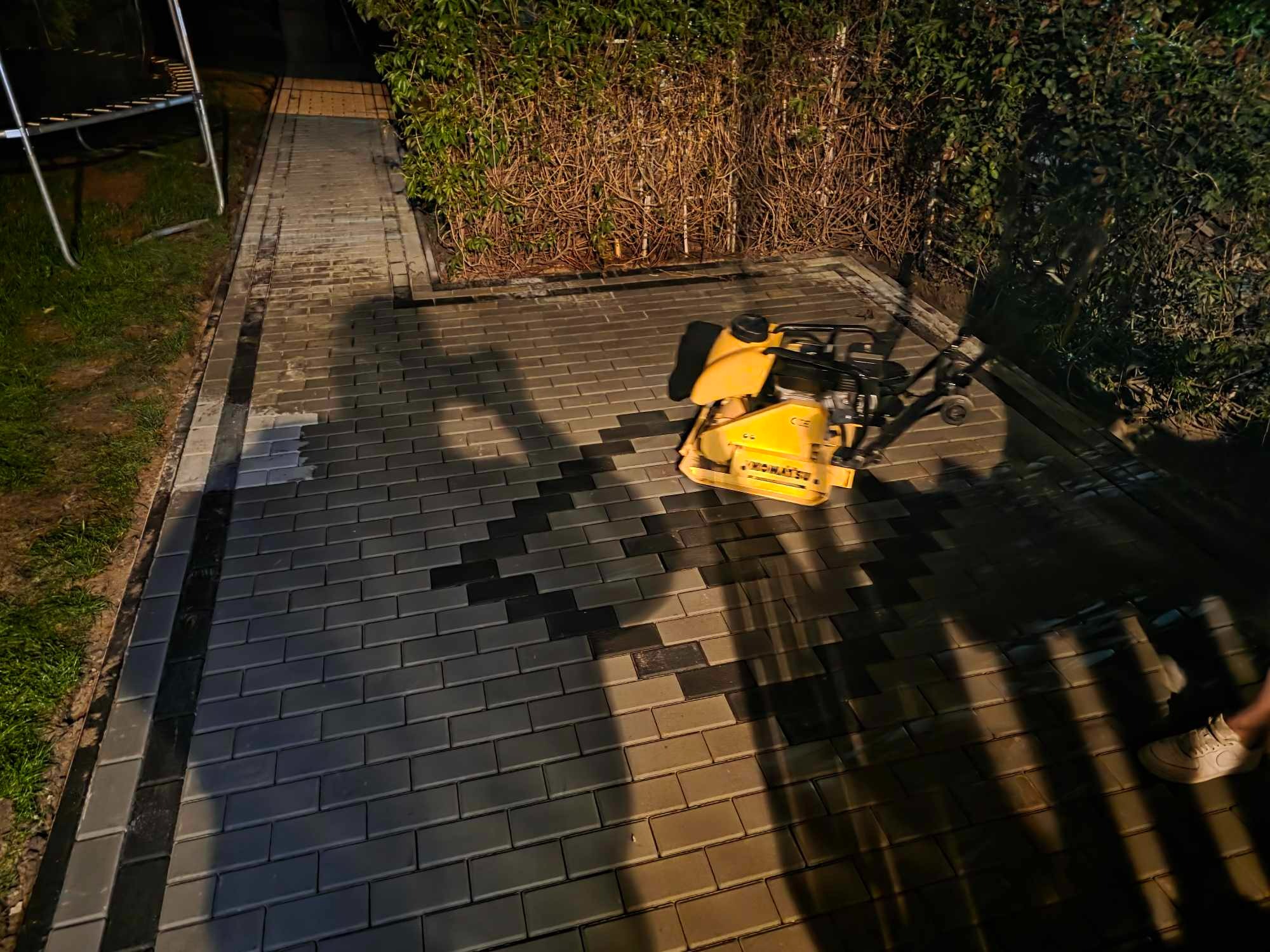 Żółta zagęszczarka Komatsu na świeżo ułożonej, szarej kostce brukowej w geometryczny wzór, oświetlonej nocą. Widoczne obrzeża i trawnik.