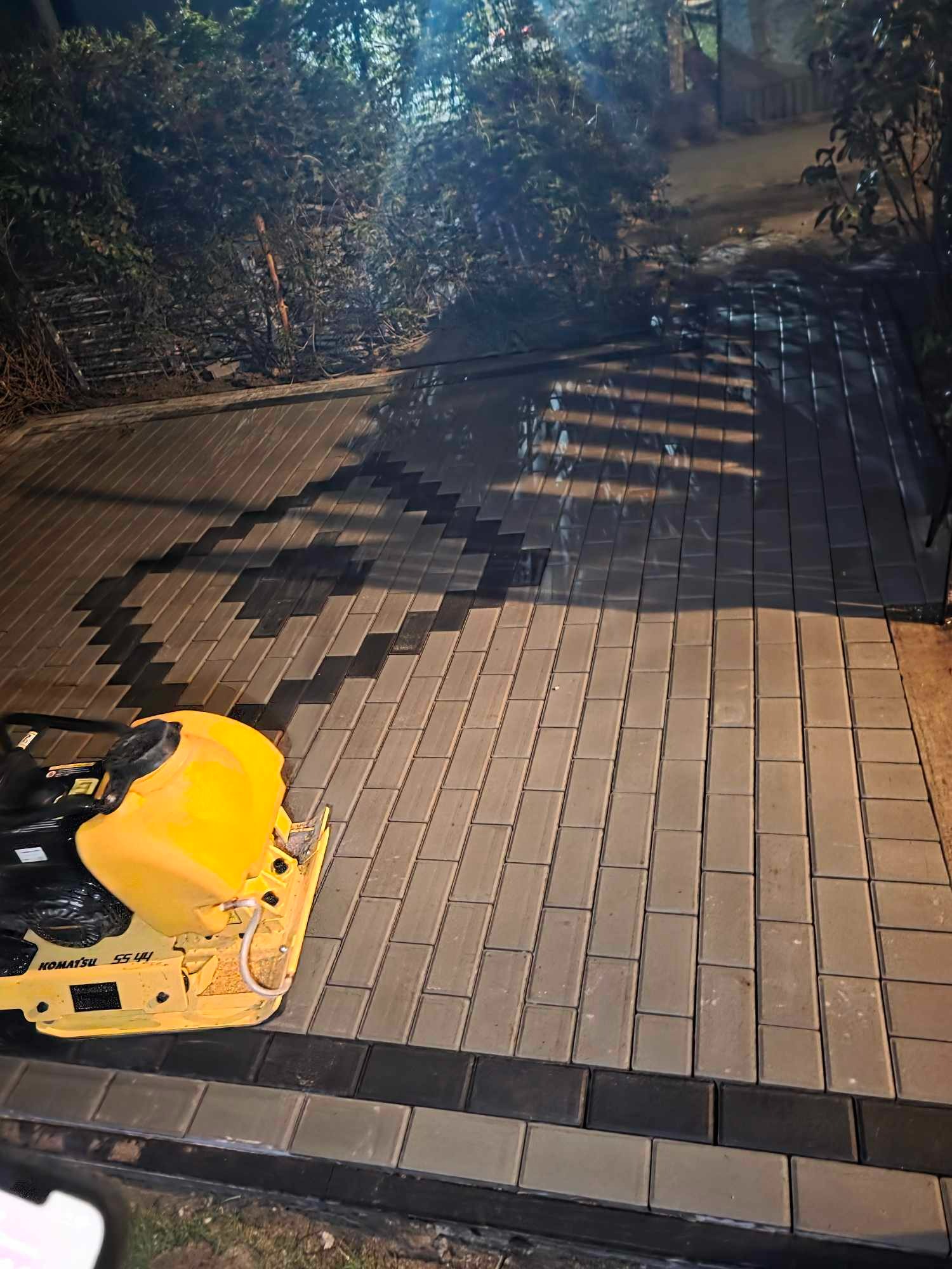 Układanie kostki brukowej z użyciem zagęszczarki Komatsu. Widoczne ułożone kostki w dwóch kolorach, tworzące wzór. Oświetlenie nocne.
