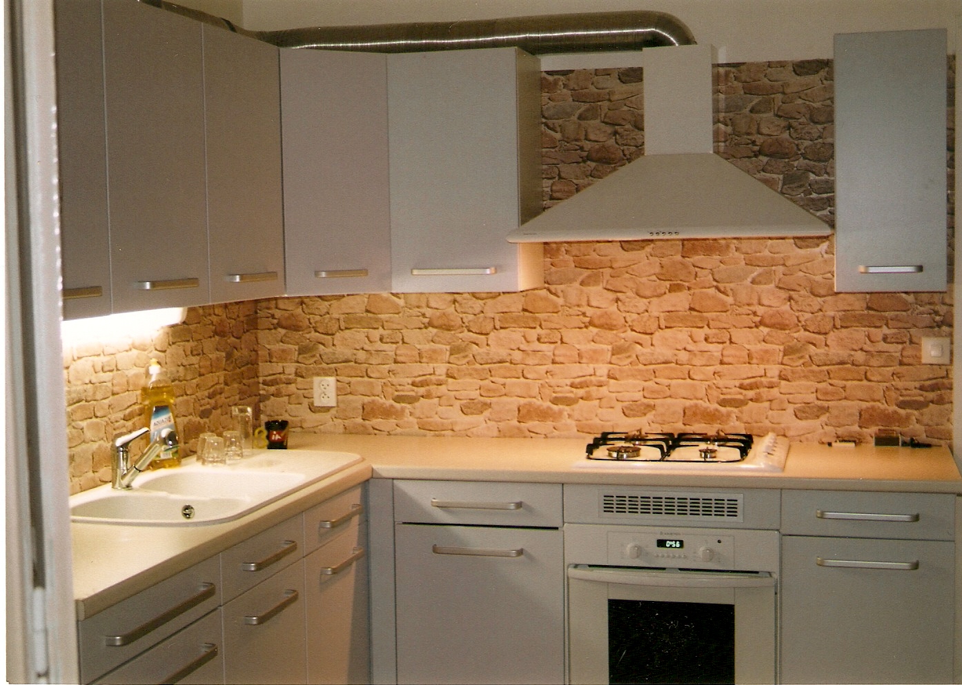 Kuchnia z szarymi szafkami, blatem i okładziną ściany imitującą kamień, oświetlona lampkami podszafkowymi, z kuchenką gazową i okapem.