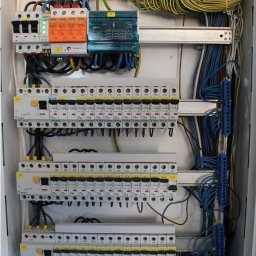 DAWID STASZEWSKI CoolPower - Rozdzielnia elektryczna z zainstalowanymi bezpiecznikami, ogranicznikami przepięć i uporządkowanymi przewodami wewnątrz białej obudowy.