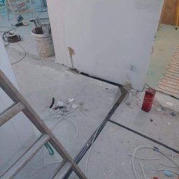 DAWID STASZEWSKI CoolPower - Wnętrze w trakcie prac elektrycznych: kable w podłodze, puszki w ścianie, drabina, narzędzia. Widoczne elementy instalacji ogrzewania podłogowego.