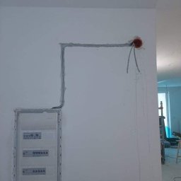 DAWID STASZEWSKI CoolPower - Wnęka pod rozdzielnicę i bruzdy pod kable elektryczne w ścianie, przygotowane do dalszych prac instalacyjnych. Widoczne przewody wychodzące z puszki.