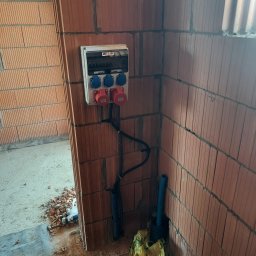 DAWID STASZEWSKI CoolPower - Rozdzielnia elektryczna z gniazdami siłowymi na ścianie z czerwonej cegły dziurawki, kable w peszlach. Instalacja w trakcie montażu w surowym stanie.